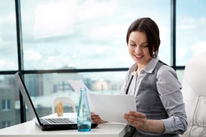 Beautiful business woman looking at papers she holding in her arms while working on computer at her office